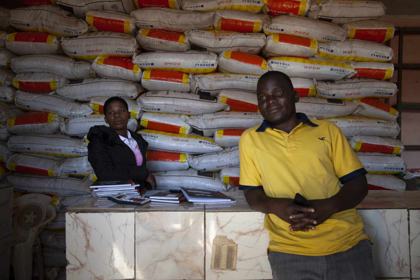 Two individuals stand in front of bags containing agricultural inputs