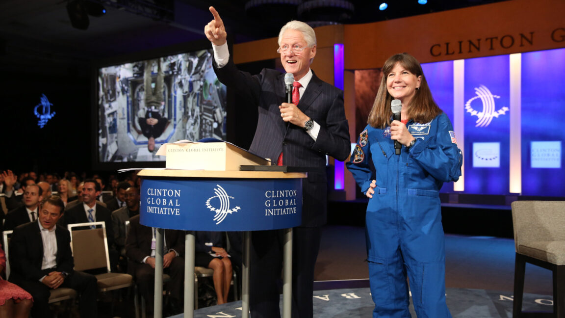 President Clinton and astronaut Cady Coleman stand onstage during a Clinton Global Initiative event. President Clinton points up at a screen. On another screen behind them is a live feed with an astronaut aboard the International Space Station.