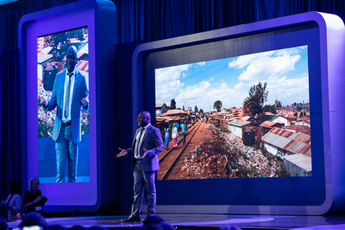 Kennedy Odede stands on stage. Behind him are large digital screens which show photos.