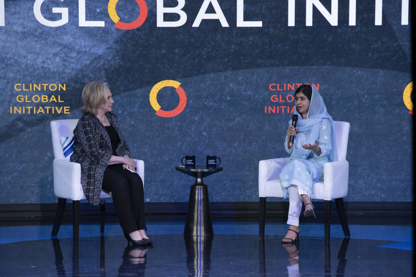 Secretary Clinton and Malala Yousafzai sit onstage. Malala speaks into a microphone.