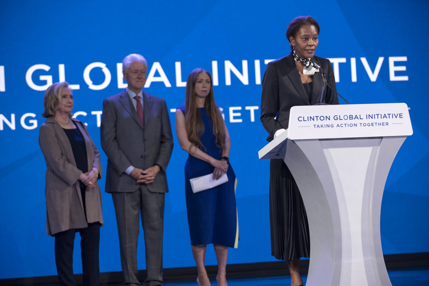 Didi Bertrand Farmer speaks onstage behind a podium. Secretary Clinton, President Clinton, and Chelsea Clinton stand behind her.