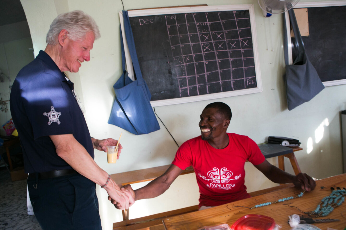 President Clinton shakes the hand of an individual putting together an artisanal piece with beads