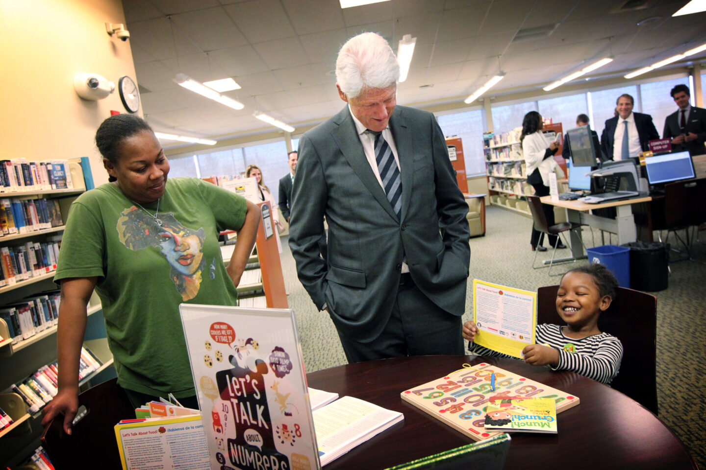President Clinton speaks with an adult and child in a library. The child holds a children's book.