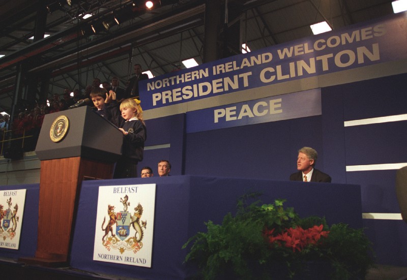 President Clinton looks on while two children speak from behind a podium. A sign behind them says: "Northern Ireland Welcomes President Clinton" and "Peace."