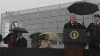 President Clinton speaks at a podium in front of the Clinton Presidential Center. It's raining and several people are holding umbrellas.