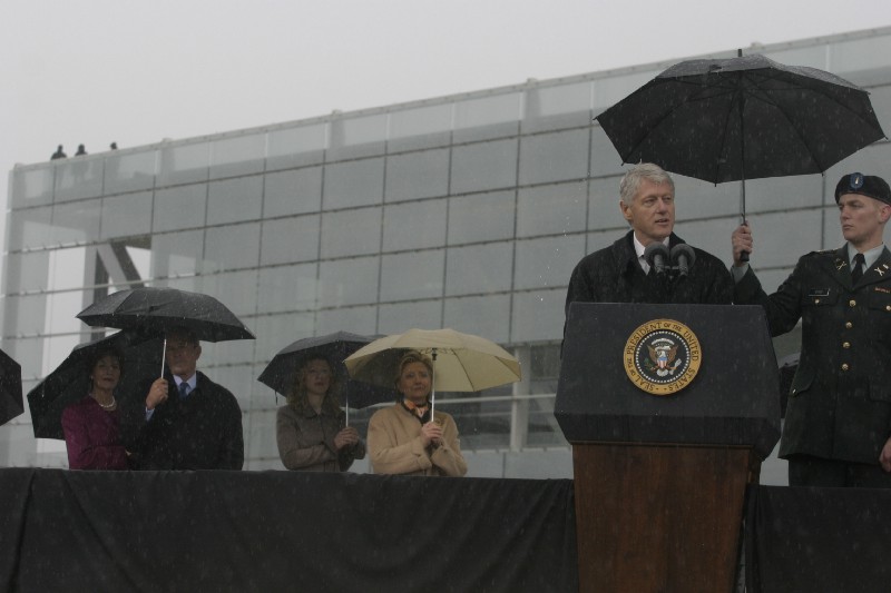 President Clinton speaks at a podium in front of the Clinton Presidential Center. It's raining and several people are holding umbrellas.