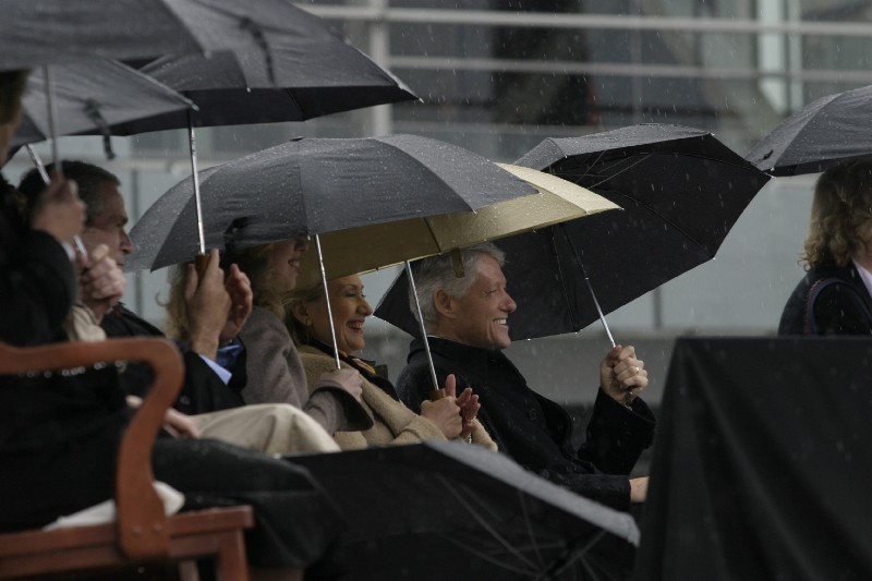 President Bill Clinton, Secretary Hillary Clinton, Chelsea Clinton, and President George W. Bush sit holding umbrellas over their heads