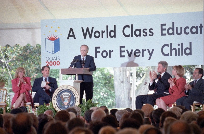 Secretary Richard Riley speaks to a large crowd from behind a podium. A sign behind him says: A World Class Education For Every Child.