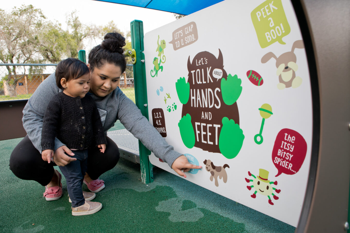 An adult and child look at a panel that says "Let's talk about hands and feet" and which contains various illustrations and prompts for reading and talking