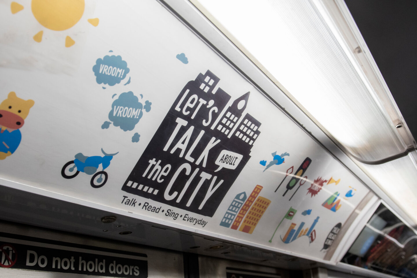 A subway ad featuring illustrations and conversations prompt. In the middle it says, "Let's talk about the city" and "Talk. Read. Sing. Everyday."