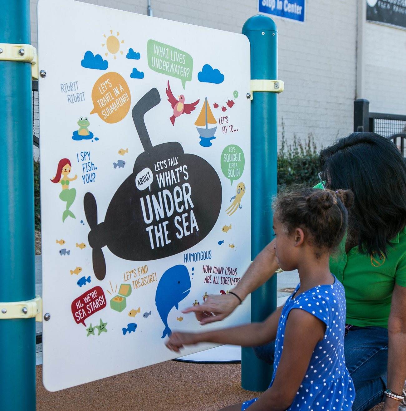 An adult and child stand outside in front of a panel with a variety of "under the sea" themed prompts for reading and talking
