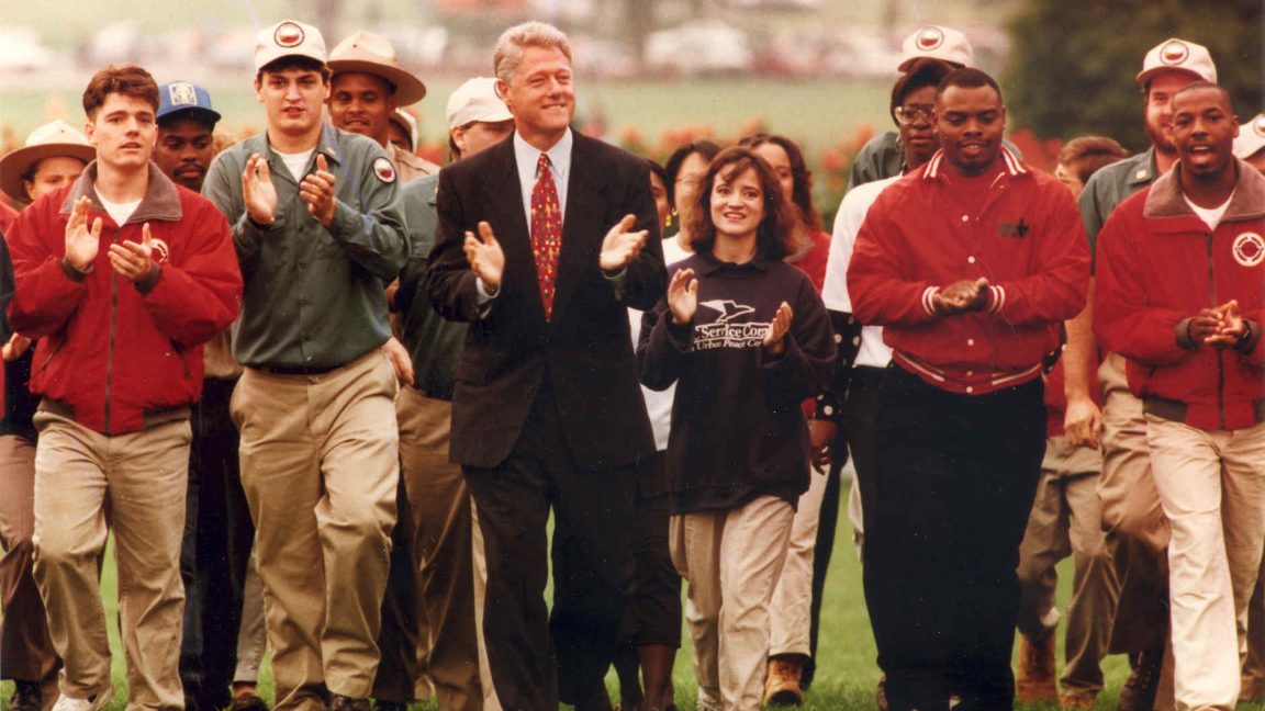 President Clinton and a group of individuals walk outside. Several are wearing red Americorps jackets.