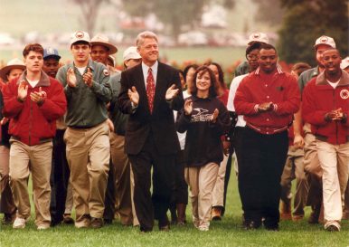 President Clinton and a group of individuals walk outside. Several are wearing red Americorps jackets.