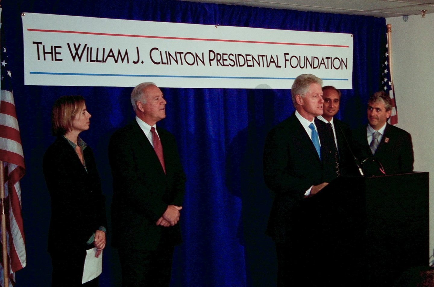 President Clinton and several individuals stand in front of a podium. A sign behind them says "The William J. Clinton Presidential Foundation"