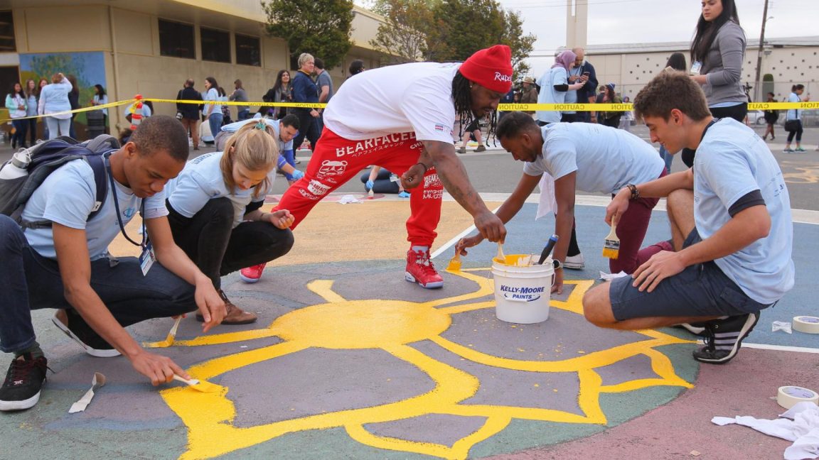 Marshawn Lynch and volunteers paint a flower design on the ground outside a school