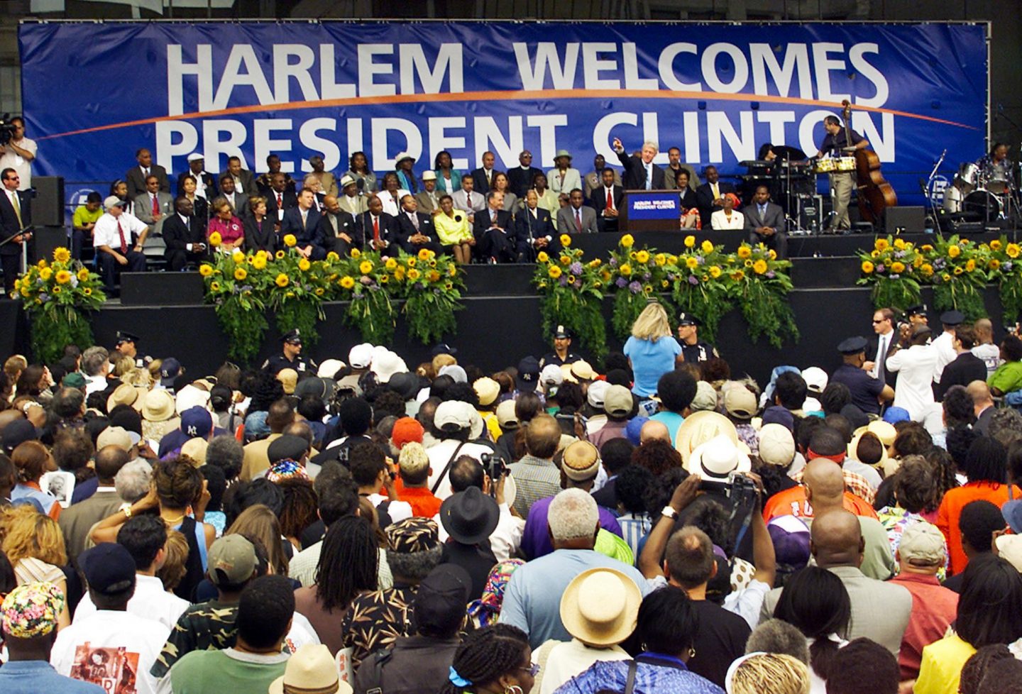 President Clinton speaks onstage in front of a large crowd. A sign behind him says: "Harlem welcomes President Clinton."