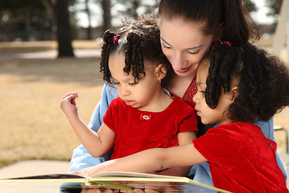 An adult reads a book outside with two children