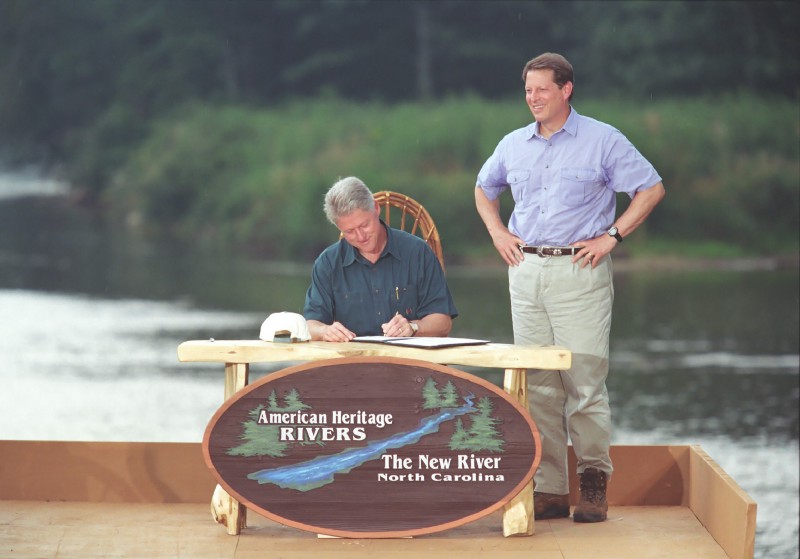 President Clinton sits at a desk placed outdoors and signs papers, with Al Gore on his left side. Behind them is a river. A podium sign reads: American Heritage Rivers. The New River. North Carolina.