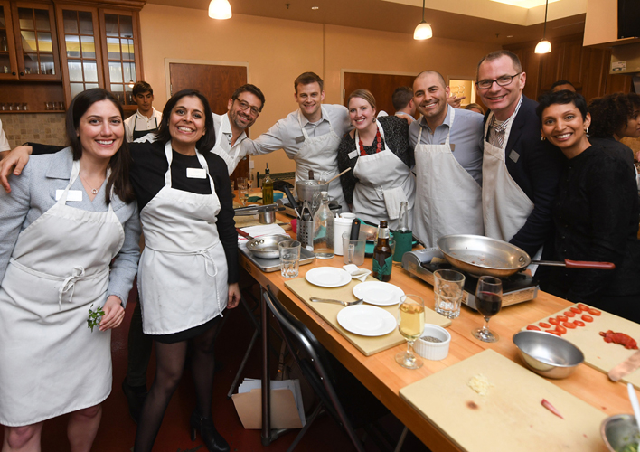 A group pictured preparing food in a kitchen. Most are wearing aprons.