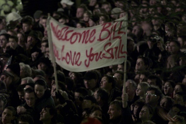 A crowd holds a sign that says: "Welcome Bill. We love you still."