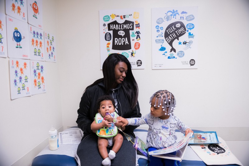 An individual sits with an infant and small child in a medical clinic. The child reads a book. On the wall behind them are posters featuring illustrations and conversation prompts. 