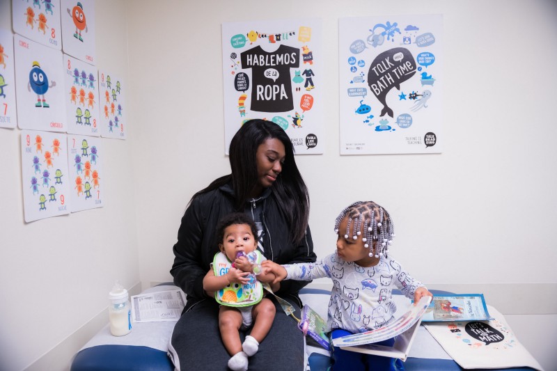 An individual sits with an infant and small child in a medical clinic. The child reads a book. On the wall behind them are posters featuring illustrations and conversation prompts.