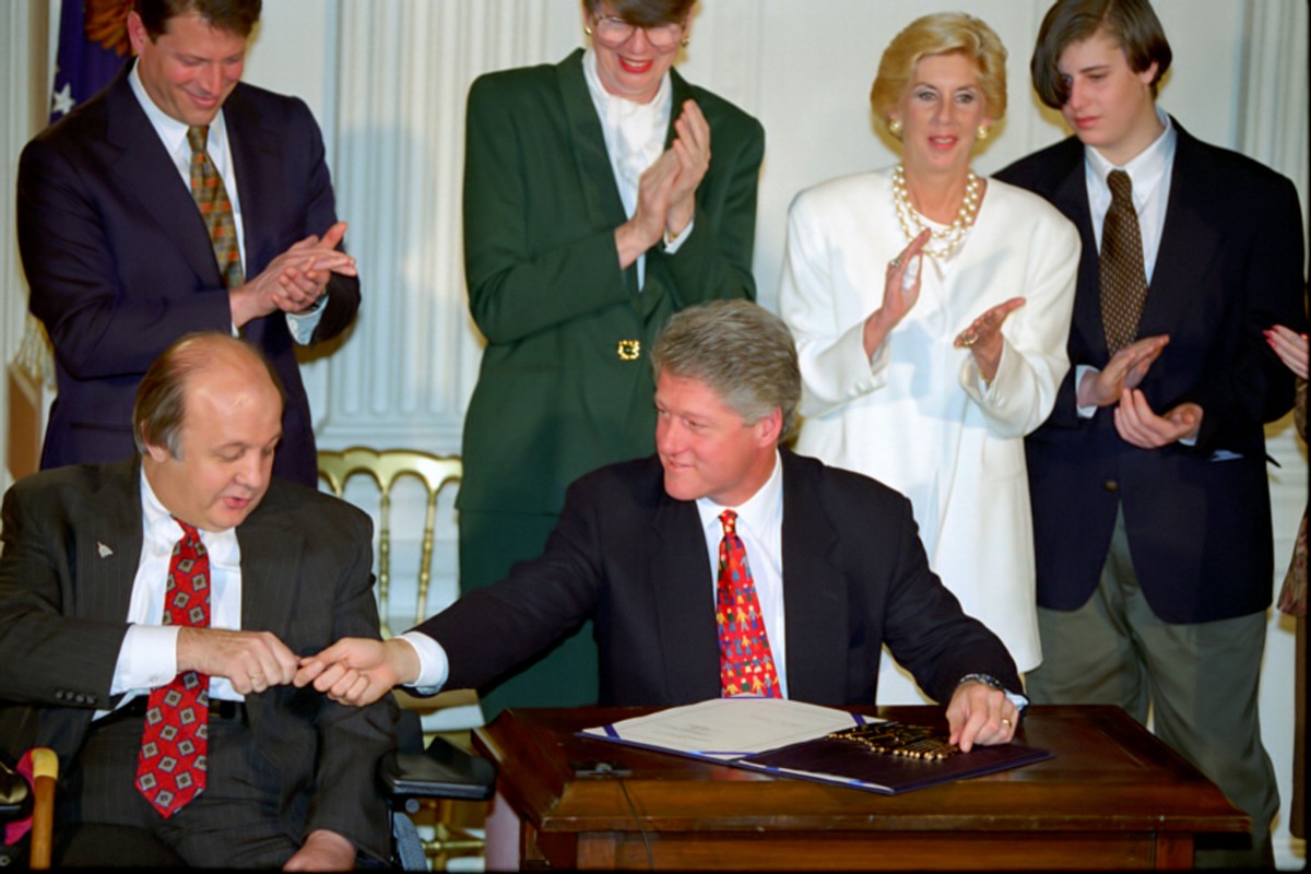 President Bill Clinton hands Jim Brady a pen at the signing of the Brady Bill. Several individuals stand behind them applauding.  