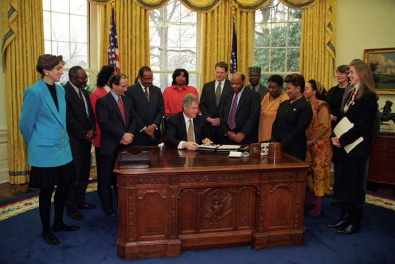 President Clinton sits at the Resolute Desk in the Oval Office and signs an executive order. A large group is gathered around. 