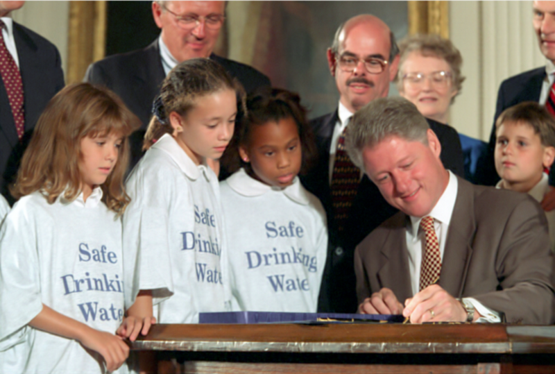 President Clinton sits at a desk while signing legislation at the White House. A large group is gathered around him, including three children wearing t-shirts that say: Safe Drinking Water.