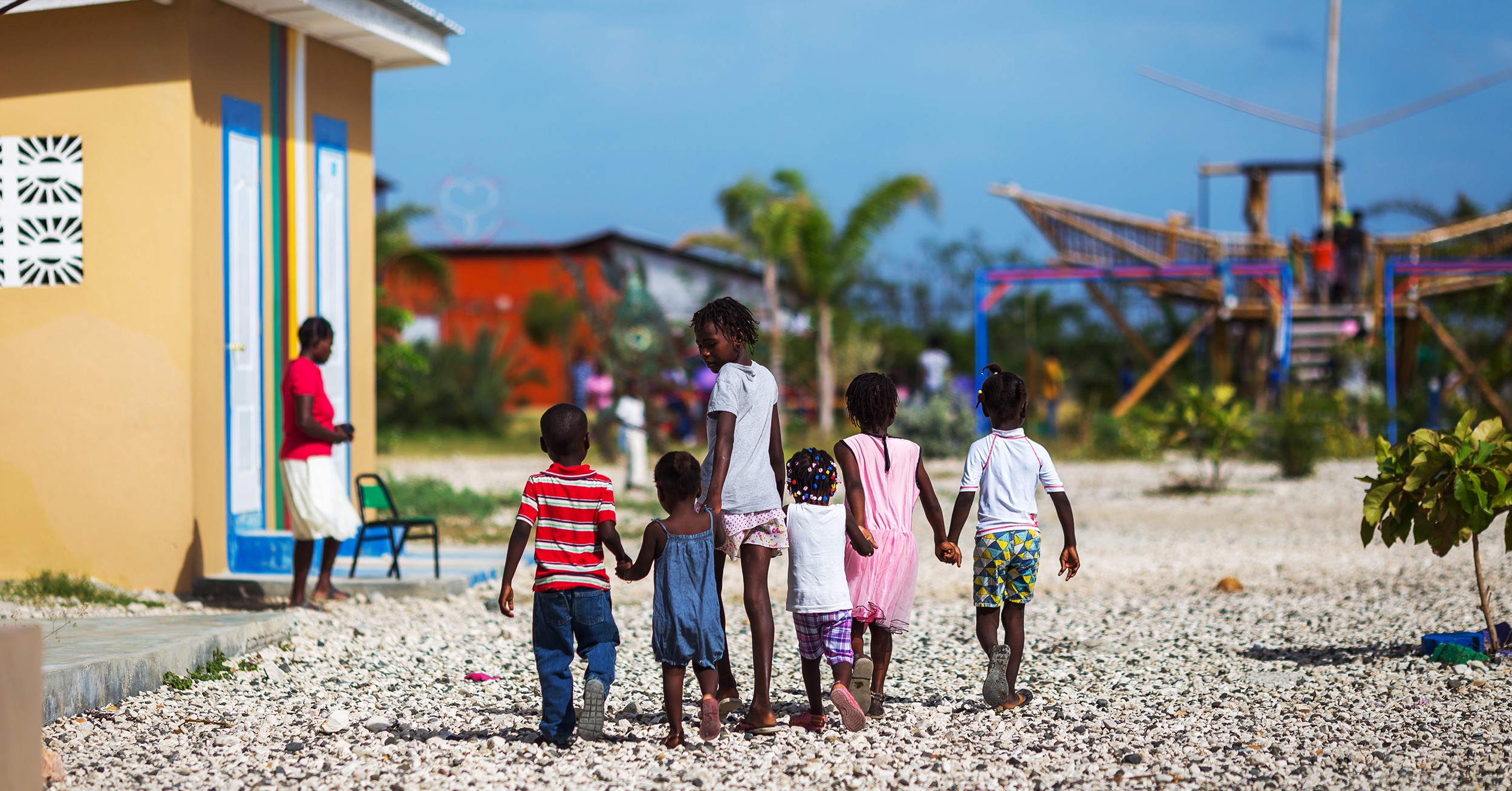 A group of children hold hands while walking. A play structure is in the background.