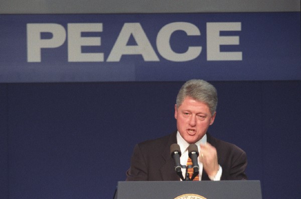 President Clinton speaks from behind a podium. A sign behind him says: Peace.