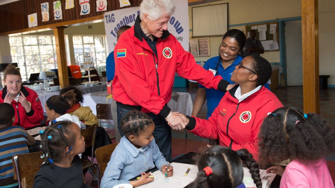 President Clinton shakes a person's hand at City Year South Africa. They both wear red City Year jackets. Nearby children sit at tables with art and reading materials.