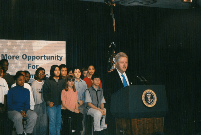 President Clinton speaks at a podium. Behind him is a group of young people.