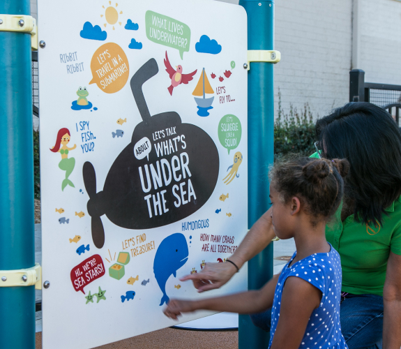 An adult and child stand outside in front of a panel with a variety of "under the sea" themed prompts for reading and talking