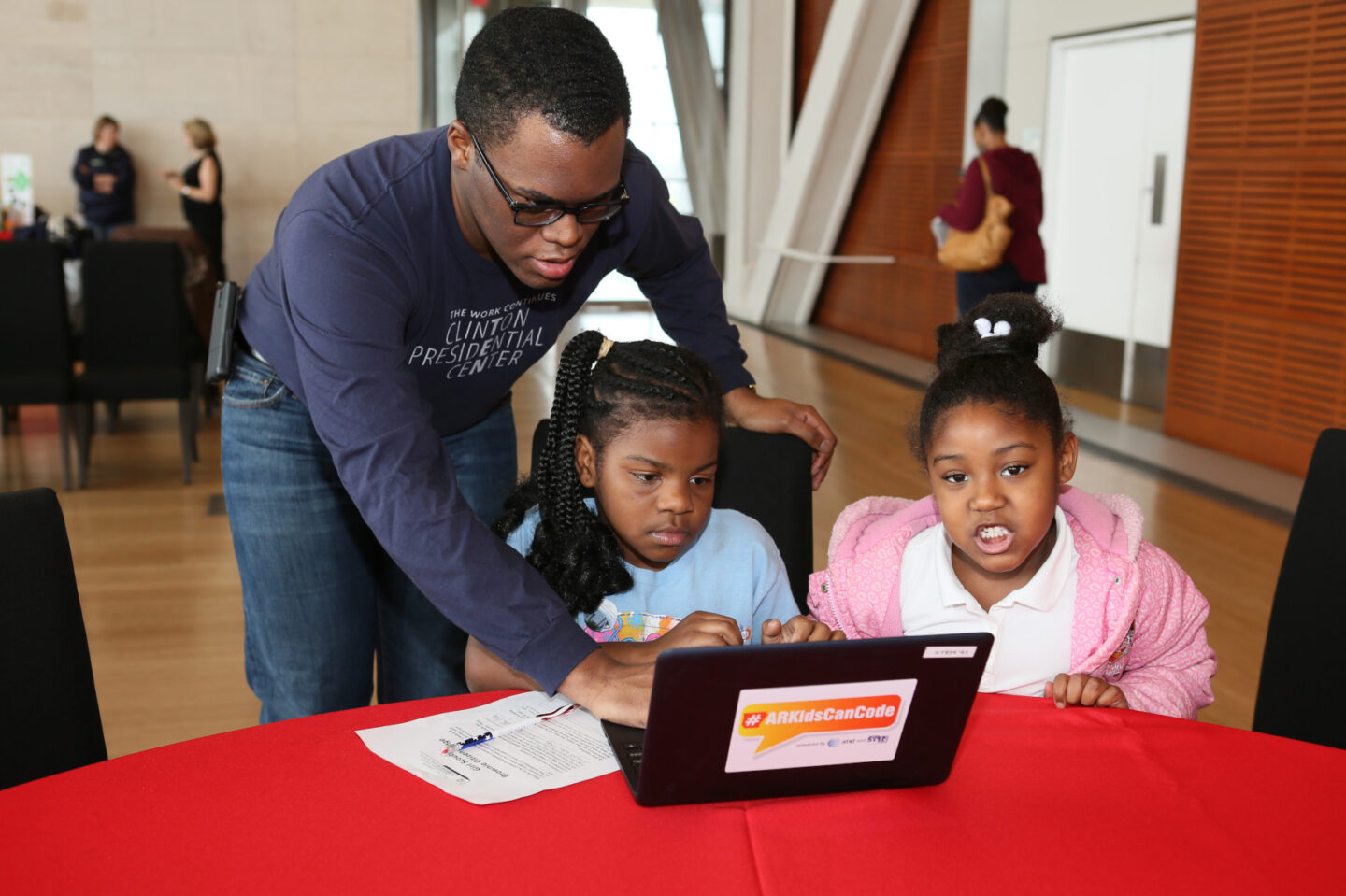 A staff member and two children use a computer with a sticker that reads #ARKidsCanCode