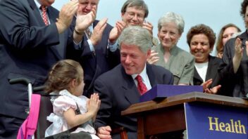 President Clinton sits behind a desk during a bill signing with a group of individuals behind him and a child seated in a wheelchair next to him.