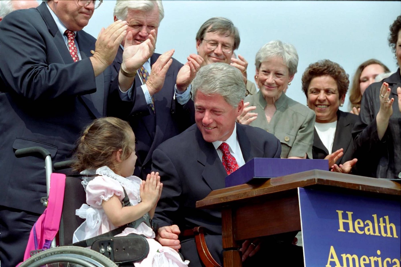 President Clinton sits behind a desk during a bill signing with a group of individuals behind him and a child seated in a wheelchair next to him.