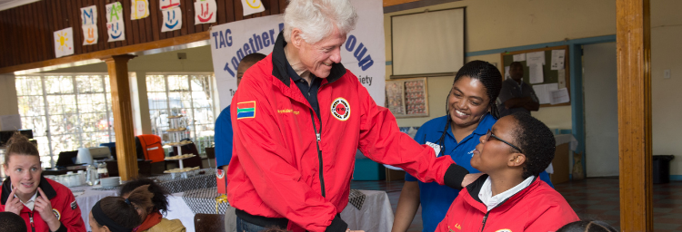 President Clinton shakes a person's hand at City Year South Africa. They both wear red City Year jackets. Nearby children sit at tables with art and reading materials.