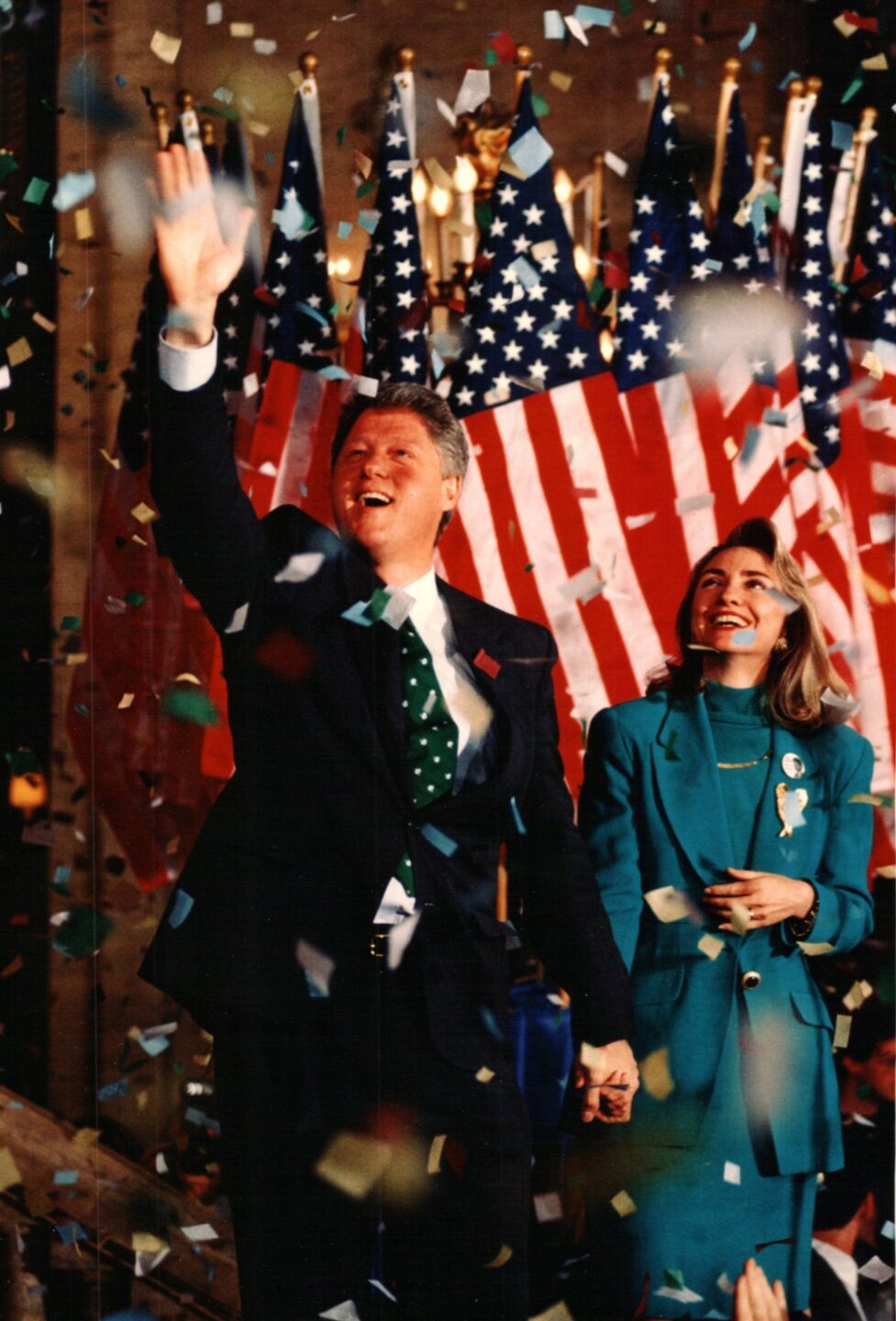 President Clinton and Secretary Clinton stand onstage during a presidential campaign rally in 1992. Confetti is falling and there are American flags behind them.