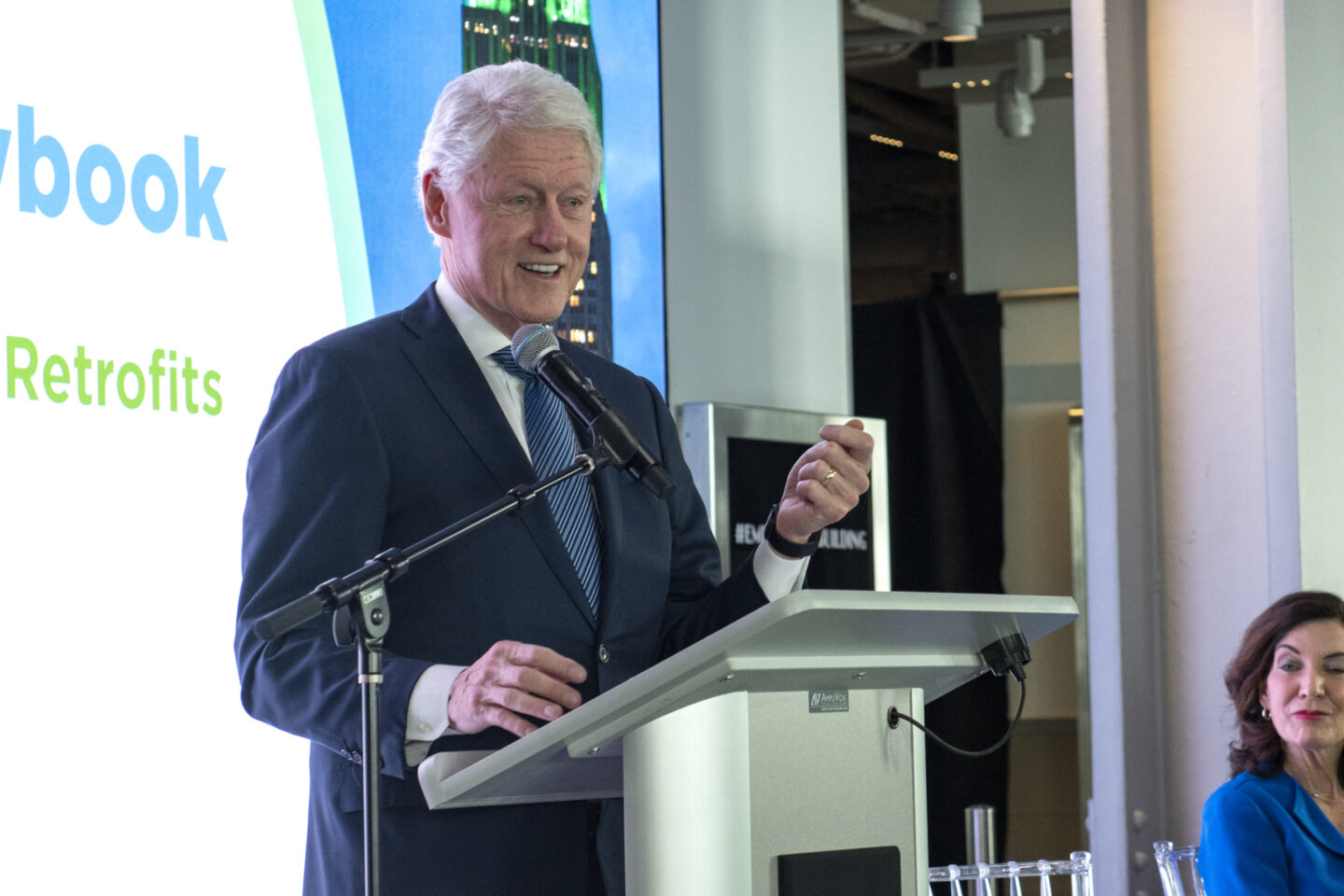 President Clinton speaks behind a lectern. Governor Kathy Hochul is seated nearby.