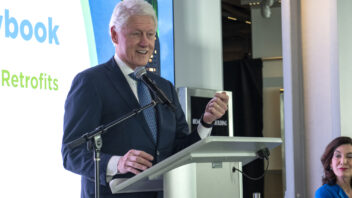 President Clinton speaks behind a lectern. Governor Kathy Hochul is seated nearby.