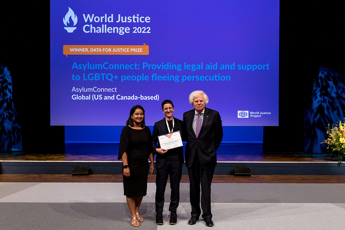 Three people stand on stage. Jamie Sgarro, center, holds an award for the Data for Justice Prize. Behind the people is a purple screen with white text reading World Justice Challenge 2022 and lists AsylumConnect as the winner of the Data for Justice Prize.