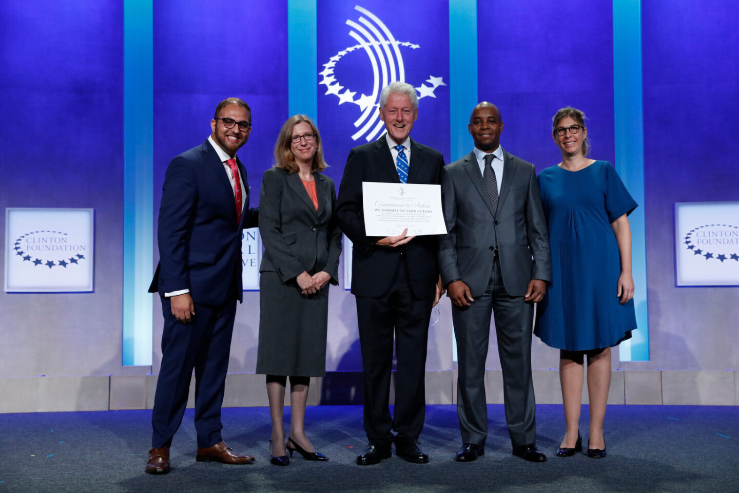 President Bill Clinton stands on a stage holding a piece of paper, with two people on either side of him.