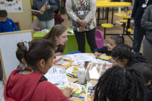 Chelsea Clinton, an adult, and several children sit at a table filled with drawing and learning materials