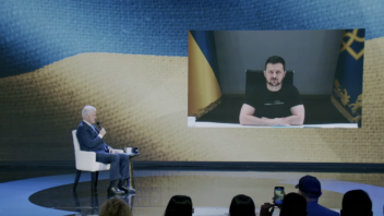 President Clinton sits onstage and President Zelenskyy appears virtually through a screen behind the stage