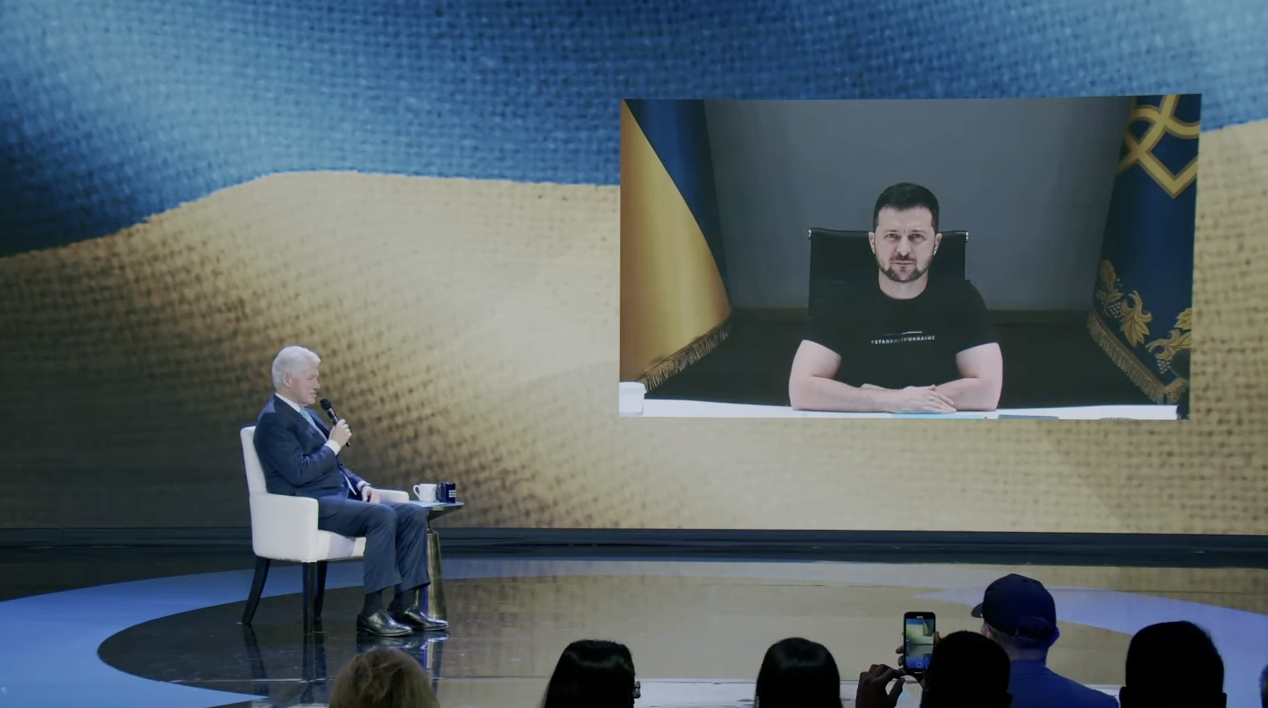 President Clinton sits onstage and President Zelenskyy appears virtually through a screen behind the stage