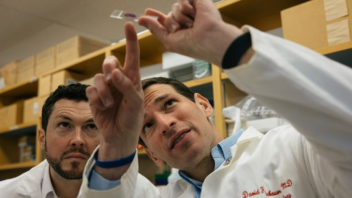 Drs. Grant Mitchell and David Fajgenbaum stand in a lab wearing white lab coats as they examine a blood specimen
